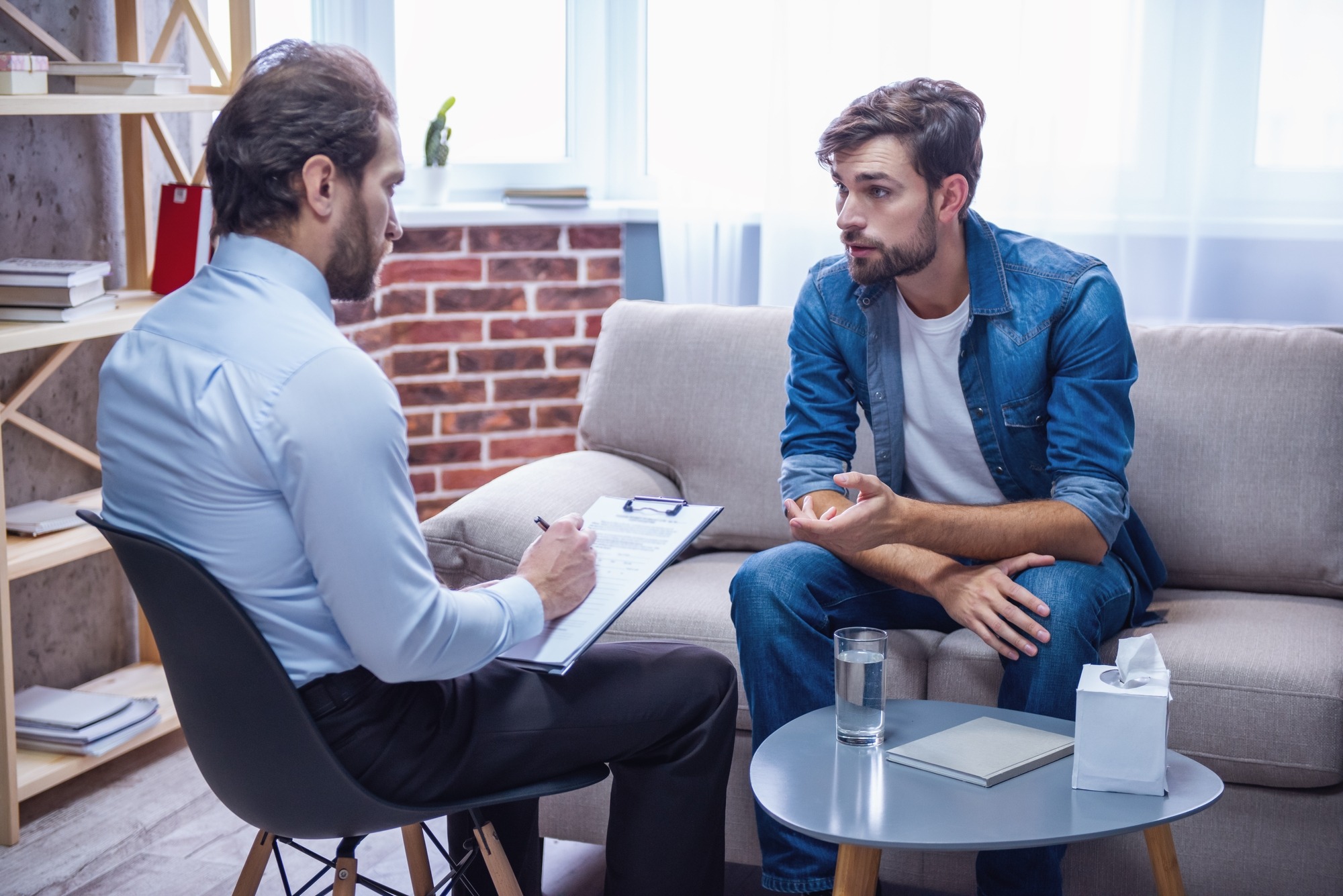 A therapist takes notes while listening to a male client discussing his thoughts in a comfortable office setting.