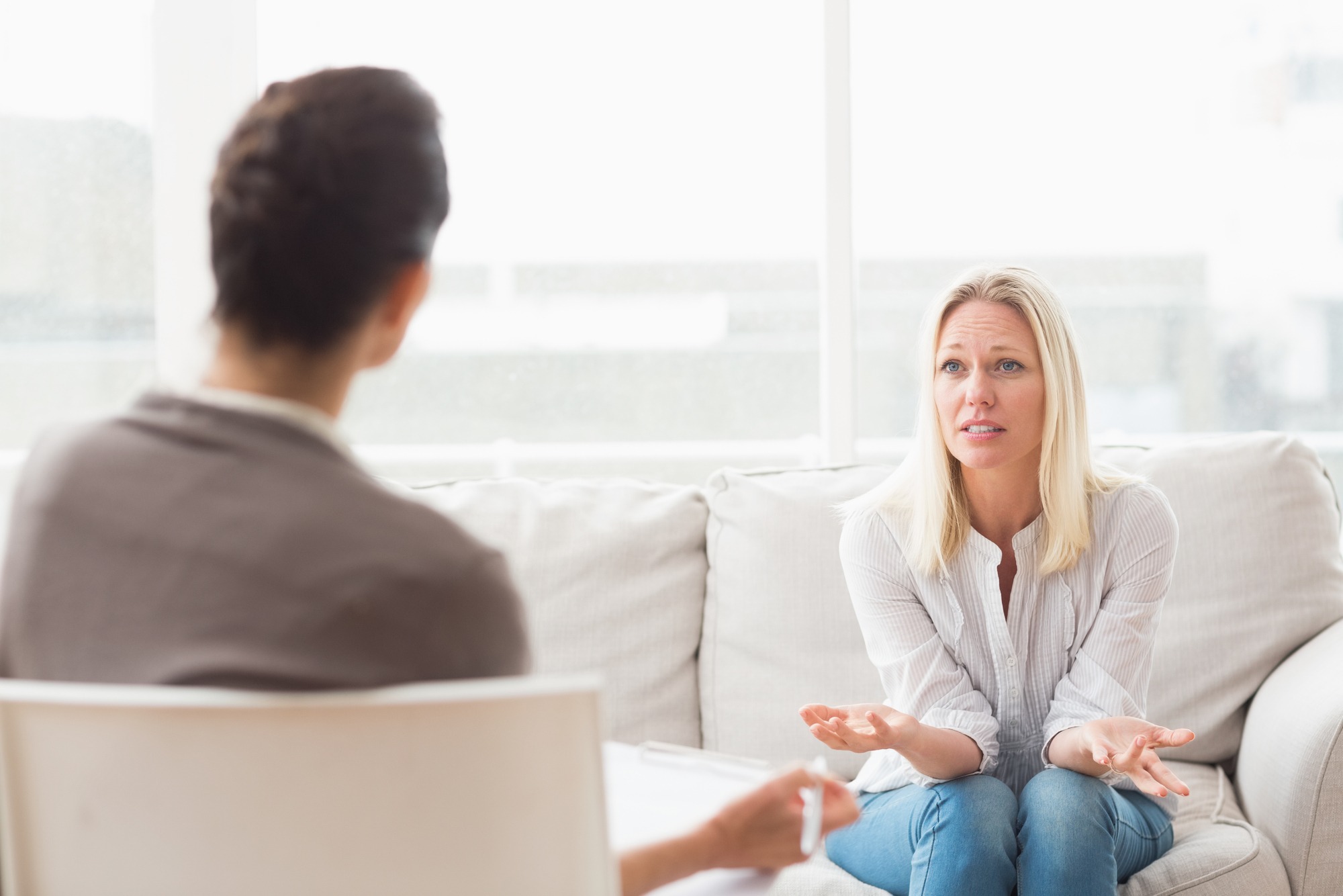 A woman with long blond hair expresses concern while seated on a couch during a therapy session. The setting is bright and open, suggesting a supportive environment.